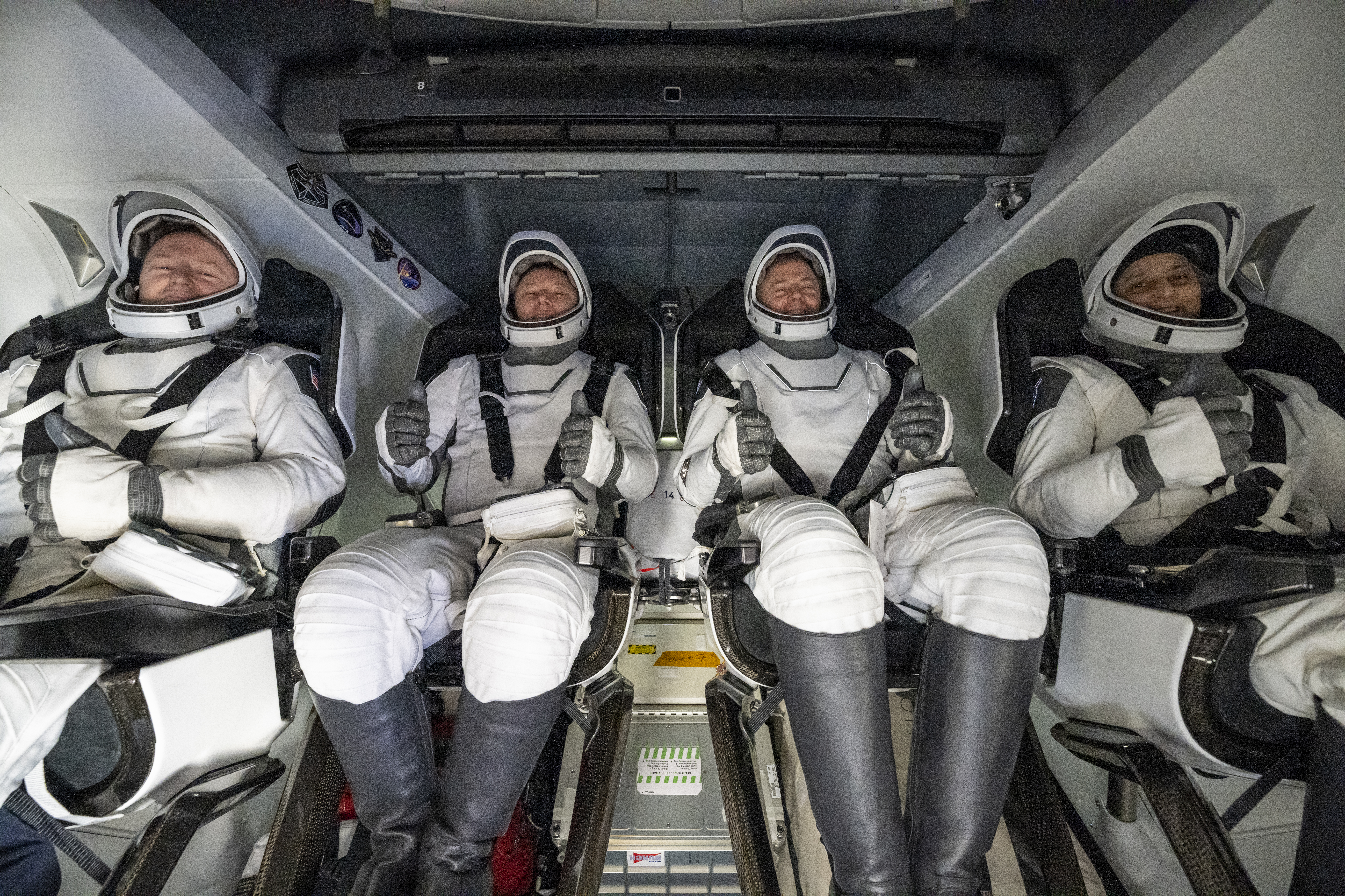 54394869292 ab31f980f5 o.jpg Four people in white spacesuits, white helmets, and black boots give a thumbs up from inside a SpaceX Dragon spacecraft.