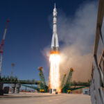 Expedition 73 Crew Heads to ISS – NASA A light gray rocket lifts off, leaving flames and light brown vapor in its wake. Green supports lie perpendicular to the rocket.