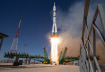 Expedition 73 Crew Heads to ISS – NASA A light gray rocket lifts off, leaving flames and light brown vapor in its wake. Green supports lie perpendicular to the rocket.