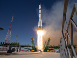 Expedition 73 Crew Heads to ISS – NASA A light gray rocket lifts off, leaving flames and light brown vapor in its wake. Green supports lie perpendicular to the rocket.