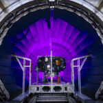 Examining NASA’s IMAP: The Interstellar Mapping and Acceleration Probe A tunnel dramatically lit in violet light dominates the photo. In front of it, a small spacecraft is on a track.
