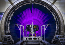 Examining NASA’s IMAP: The Interstellar Mapping and Acceleration Probe A tunnel dramatically lit in violet light dominates the photo. In front of it, a small spacecraft is on a track.