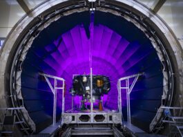 Examining NASA’s IMAP: The Interstellar Mapping and Acceleration Probe A tunnel dramatically lit in violet light dominates the photo. In front of it, a small spacecraft is on a track.