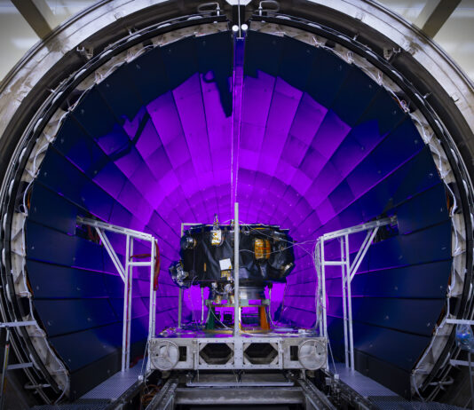 Examining NASA’s IMAP: The Interstellar Mapping and Acceleration Probe A tunnel dramatically lit in violet light dominates the photo. In front of it, a small spacecraft is on a track.