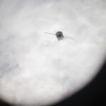 NASA Returns to Earth The Soyuz spacecraft looks like a cylinder with two lines stretching outward from it. It is oriented toward Earth, which is covered with white clouds.