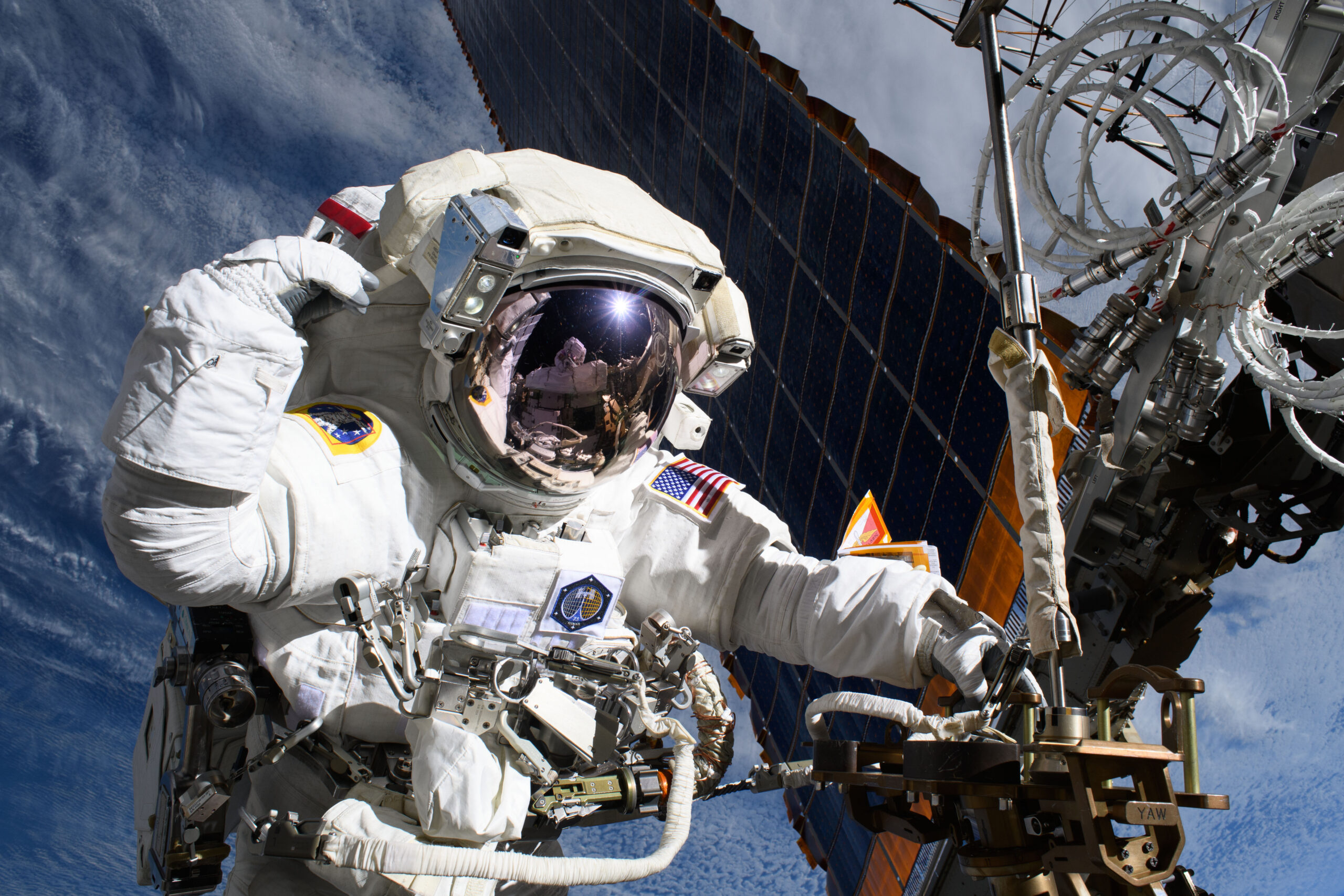54492396655 c6ba83df1d o 1.jpg An astronaut outside of the International Space Station has one hand on a truss near a solar panel. Her other hand is by her head. Reflected in her helmet is astronaut Nichole Ayers, also in a white spacesuit, taking the photo. Earth