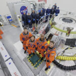 Artemis II Astronauts Undergo Training in Orion Capsule The Artemis II crew stands in front of the Orion spacecraft. They are wearing orange spacesuits without helmets. Orion is silver-colored and reflective. There is a round railing surrounding the spacecraft. There is also a group of people wearing blue jumpsuits, hairnets, and gloves standing behind Orion; they are members of the Artemis II closeout crew.