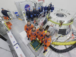 Artemis II Astronauts Undergo Training in Orion Capsule The Artemis II crew stands in front of the Orion spacecraft. They are wearing orange spacesuits without helmets. Orion is silver-colored and reflective. There is a round railing surrounding the spacecraft. There is also a group of people wearing blue jumpsuits, hairnets, and gloves standing behind Orion; they are members of the Artemis II closeout crew.