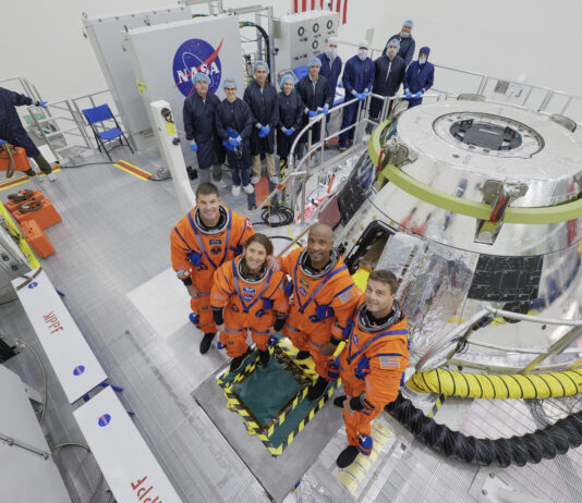 Artemis II Astronauts Undergo Training in Orion Capsule The Artemis II crew stands in front of the Orion spacecraft. They are wearing orange spacesuits without helmets. Orion is silver-colored and reflective. There is a round railing surrounding the spacecraft. There is also a group of people wearing blue jumpsuits, hairnets, and gloves standing behind Orion; they are members of the Artemis II closeout crew.