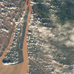 NASA Explores Southern Vietnam’s Bassac River A view of a brown river and an islet from the International Space Station. The land visible on the right is mostly dark green, while the land on the left is brown with green edges along the river. Clumps of smaller white clouds dot the photo, almost like lines of dots. Wispier clouds are visible along the right side of the image.