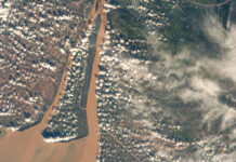 NASA Explores Southern Vietnam’s Bassac River A view of a brown river and an islet from the International Space Station. The land visible on the right is mostly dark green, while the land on the left is brown with green edges along the river. Clumps of smaller white clouds dot the photo, almost like lines of dots. Wispier clouds are visible along the right side of the image.
