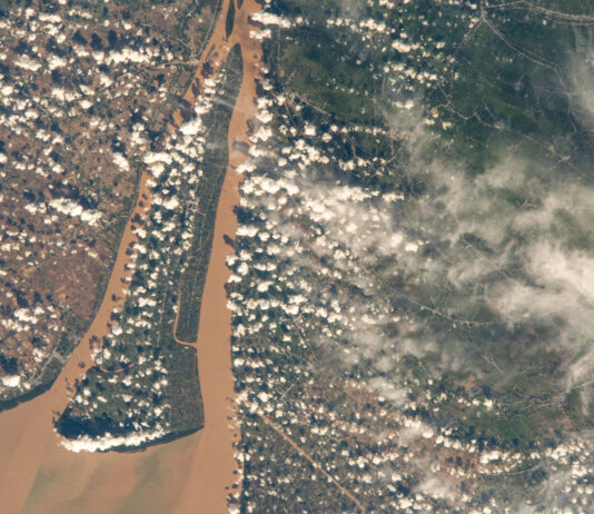 NASA Explores Southern Vietnam’s Bassac River A view of a brown river and an islet from the International Space Station. The land visible on the right is mostly dark green, while the land on the left is brown with green edges along the river. Clumps of smaller white clouds dot the photo, almost like lines of dots. Wispier clouds are visible along the right side of the image.