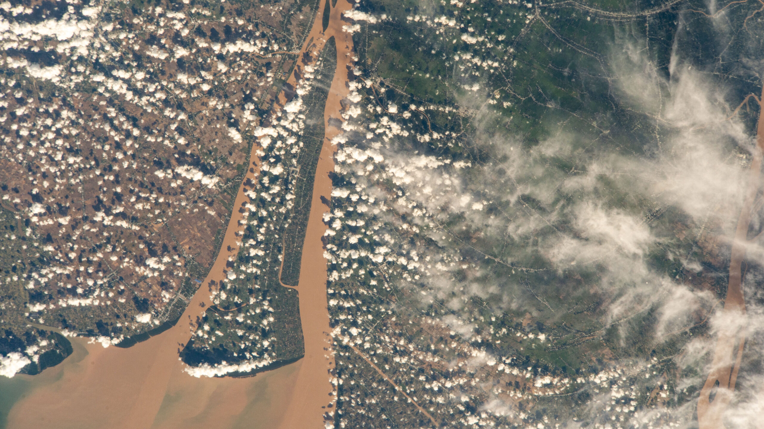 54949698099 88d64018a7 o.jpg A view of a brown river and an islet from the International Space Station. The land visible on the right is mostly dark green, while the land on the left is brown with green edges along the river. Clumps of smaller white clouds dot the photo, almost like lines of dots. Wispier clouds are visible along the right side of the image.