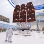 NASA Finalizes Nancy Grace Roman Space Telescope Construction Two people in white jumpsuits look up at a large telescope with three black and orange panels facing them. They are in a large white cleanroom with equipment and scaffolding.