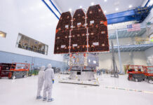 NASA Finalizes Nancy Grace Roman Space Telescope Construction Two people in white jumpsuits look up at a large telescope with three black and orange panels facing them. They are in a large white cleanroom with equipment and scaffolding.