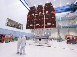 NASA Finalizes Nancy Grace Roman Space Telescope Construction Two people in white jumpsuits look up at a large telescope with three black and orange panels facing them. They are in a large white cleanroom with equipment and scaffolding.