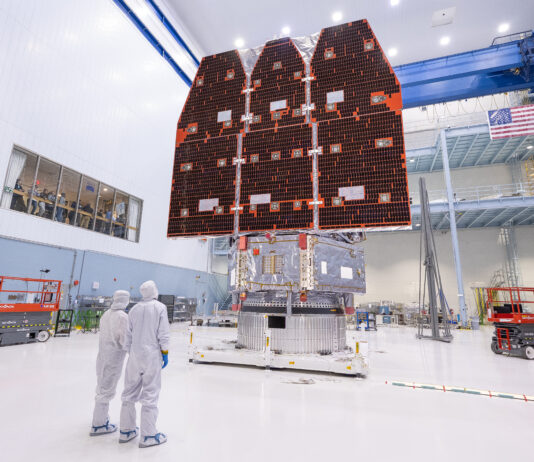 NASA Finalizes Nancy Grace Roman Space Telescope Construction Two people in white jumpsuits look up at a large telescope with three black and orange panels facing them. They are in a large white cleanroom with equipment and scaffolding.