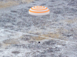 Astronaut Jonny Kim Completes Mission, Arrives Back on Earth An orange and white striped parachute attached to a black spacecraft. The spacecraft is very small compared to the parachute. It is landing on ground that has white frost on it.