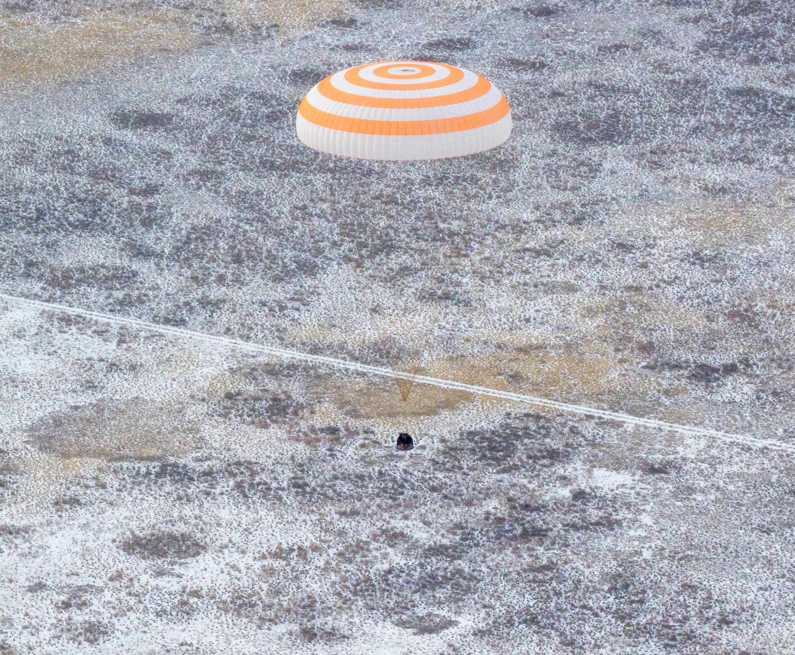 54972913396 65ce210d10 o.jpg An orange and white striped parachute attached to a black spacecraft. The spacecraft is very small compared to the parachute. It is landing on ground that has white frost on it.