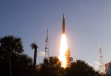 Artemis II Crew Launches to Moon with NASA Mission The Artemis II Moon rocket lifts off. There is a bright yellow-orange plume of fire underneath the rocket. The trees in the foreground appear in shadow.