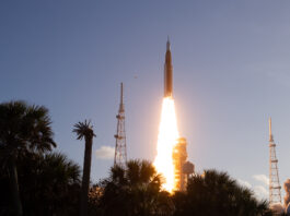 Artemis II Crew Launches to Moon with NASA Mission The Artemis II Moon rocket lifts off. There is a bright yellow-orange plume of fire underneath the rocket. The trees in the foreground appear in shadow.
