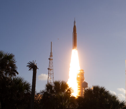 Artemis II Crew Launches to Moon with NASA Mission The Artemis II Moon rocket lifts off. There is a bright yellow-orange plume of fire underneath the rocket. The trees in the foreground appear in shadow.