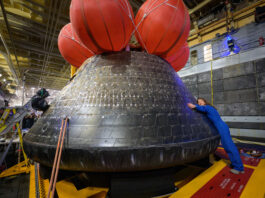 NASA Unveils New Initiative for Comfort in Space Living NASA astronaut Christina Koch hugs the Orion spacecraft with her face toward the camera. She is wearing a bright blue jumpsuit. Orion is a rounded cone shaped spacecraft. Its exterior is a sooty black. There are several large orange balloons attached to its top. Orion looks about twice Koch