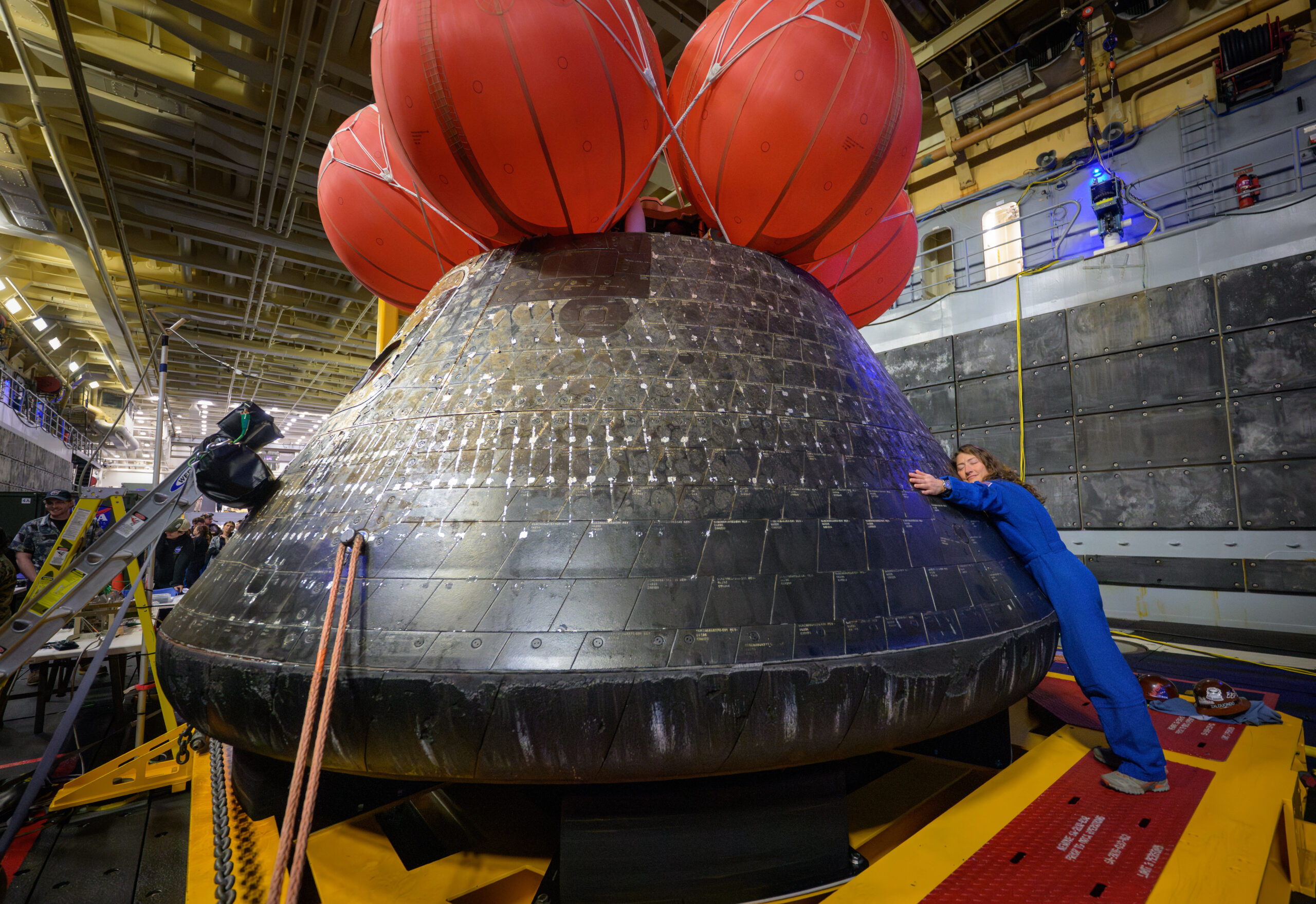 NASA astronaut Christina Koch hugs the Orion spacecraft with her face toward the camera. She is wearing a bright blue jumpsuit. Orion is a rounded cone shaped spacecraft. Its exterior is a sooty black. There are several large orange balloons attached to its top. Orion looks about twice Koch