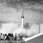 Cape Canaveral Hosts Inaugural NASA Rocket Launch In the foreground, four people stand outside near equipment as a white rocket launches in front of them. White vapor clouds cover the ground and much of the scaffolding at the launch site. The photo is black and white.