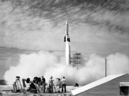 Cape Canaveral Hosts Inaugural NASA Rocket Launch In the foreground, four people stand outside near equipment as a white rocket launches in front of them. White vapor clouds cover the ground and much of the scaffolding at the launch site. The photo is black and white.