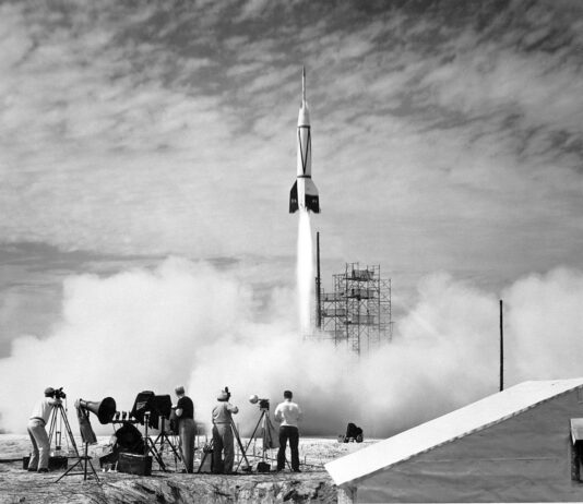 Cape Canaveral Hosts Inaugural NASA Rocket Launch In the foreground, four people stand outside near equipment as a white rocket launches in front of them. White vapor clouds cover the ground and much of the scaffolding at the launch site. The photo is black and white.