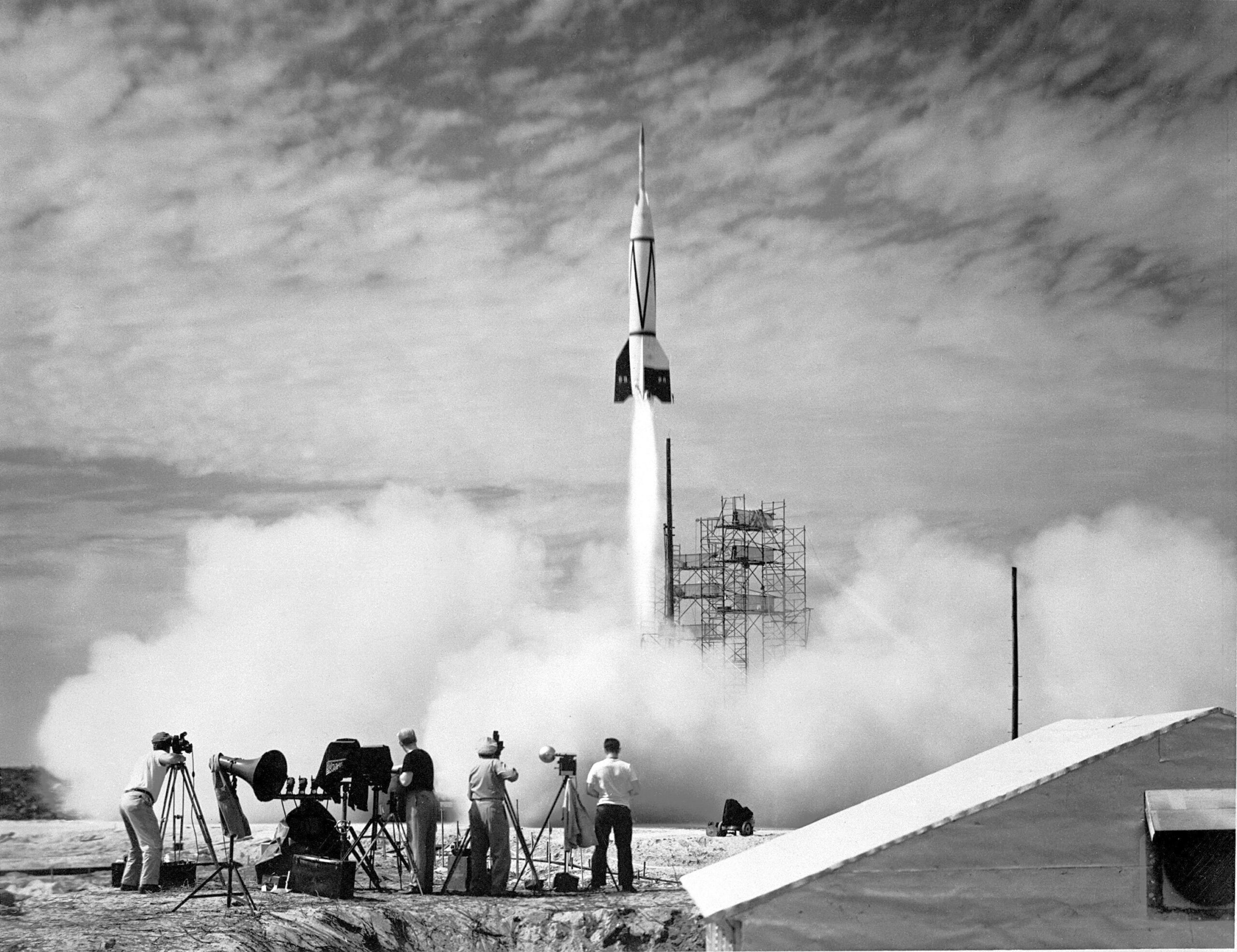 66p 0631orig.jpg In the foreground, four people stand outside near equipment as a white rocket launches in front of them. White vapor clouds cover the ground and much of the scaffolding at the launch site. The photo is black and white.