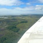 NASA Planes Conduct Science Missions for Air Quality Data The image shows an aerial view of the Chesapeake Bay, with groves of trees, rivers, a body of water in the distance, and green land. The horizon, a third of the way down the image, separates the blue cloudy sky from the land. On the right of the image is a white wing of a plane.
