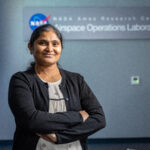 NASA Database Expert Achieves Her Dream Role A woman wearing a white blouse with small black patterns on it and a black cardigan stands smiling with her arms crossed, in front of a sign reading