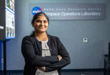 NASA Database Expert Achieves Her Dream Role A woman wearing a white blouse with small black patterns on it and a black cardigan stands smiling with her arms crossed, in front of a sign reading