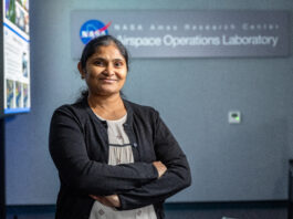 NASA Database Expert Achieves Her Dream Role A woman wearing a white blouse with small black patterns on it and a black cardigan stands smiling with her arms crossed, in front of a sign reading