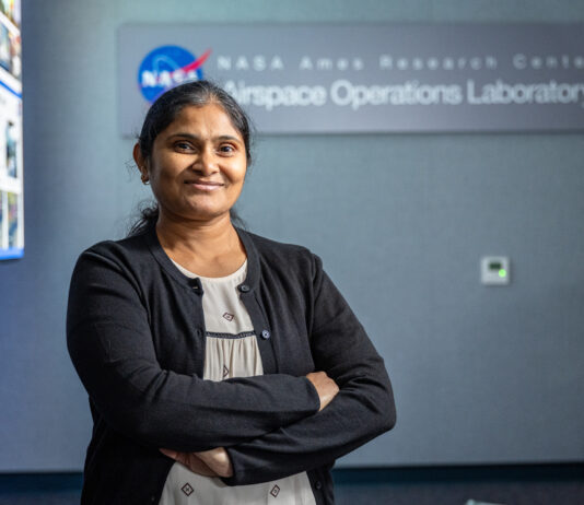 NASA Database Expert Achieves Her Dream Role A woman wearing a white blouse with small black patterns on it and a black cardigan stands smiling with her arms crossed, in front of a sign reading