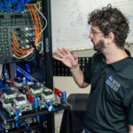 Understanding NASA’s Distributed Spacecraft Autonomy A man stands in front of a computer server and gestures towards the racks and cables.