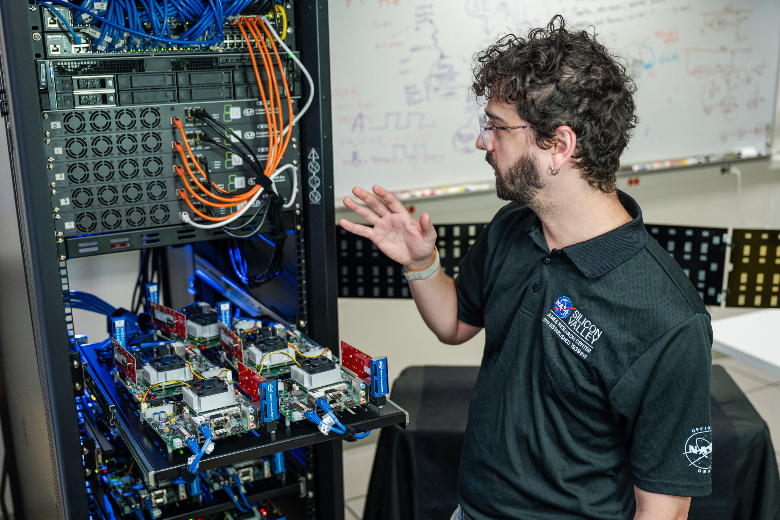 acd24 0190 018.jpg A man stands in front of a computer server and gestures towards the racks and cables.
