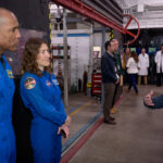 Artemis Crew and Orion Leaders Tour NASA Ames Facility Two astronauts in blue jumpsuits stand at the left of the image, listening to a man speaking on the right side of the image. People and equipment fill the background.