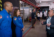 Artemis Crew and Orion Leaders Tour NASA Ames Facility Two astronauts in blue jumpsuits stand at the left of the image, listening to a man speaking on the right side of the image. People and equipment fill the background.