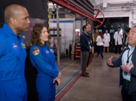 Artemis Crew and Orion Leaders Tour NASA Ames Facility Two astronauts in blue jumpsuits stand at the left of the image, listening to a man speaking on the right side of the image. People and equipment fill the background.