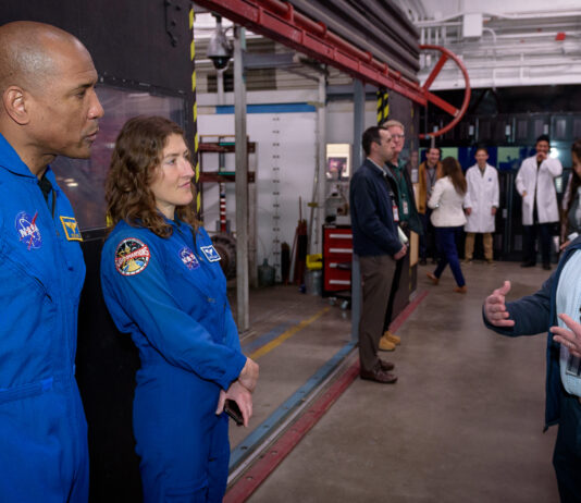 Artemis Crew and Orion Leaders Tour NASA Ames Facility Two astronauts in blue jumpsuits stand at the left of the image, listening to a man speaking on the right side of the image. People and equipment fill the background.