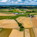 NASA-ISRO Collaboration to Track Crop Growth from Planting to Harvest Aerial view of Amish countryside with patchwork fields in shades of tan, green, and brown. White farmhouses and barns dot the landscape under a blue sky with fluffy white clouds.