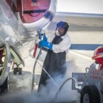 NASA’s ER-2 Aircraft Collects Crucial Atmospheric Data A man clad in a white protective suit, black apron, and blue gloves administers liquid oxygen from a tank to a parked aircraft via hose. Gas clouds hover near his feet and the man is wearing a helmet with a clear visor over his face.
