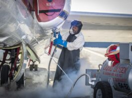 NASA’s ER-2 Aircraft Collects Crucial Atmospheric Data A man clad in a white protective suit, black apron, and blue gloves administers liquid oxygen from a tank to a parked aircraft via hose. Gas clouds hover near his feet and the man is wearing a helmet with a clear visor over his face.