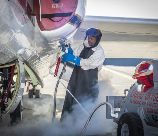NASA’s ER-2 Aircraft Collects Crucial Atmospheric Data A man clad in a white protective suit, black apron, and blue gloves administers liquid oxygen from a tank to a parked aircraft via hose. Gas clouds hover near his feet and the man is wearing a helmet with a clear visor over his face.