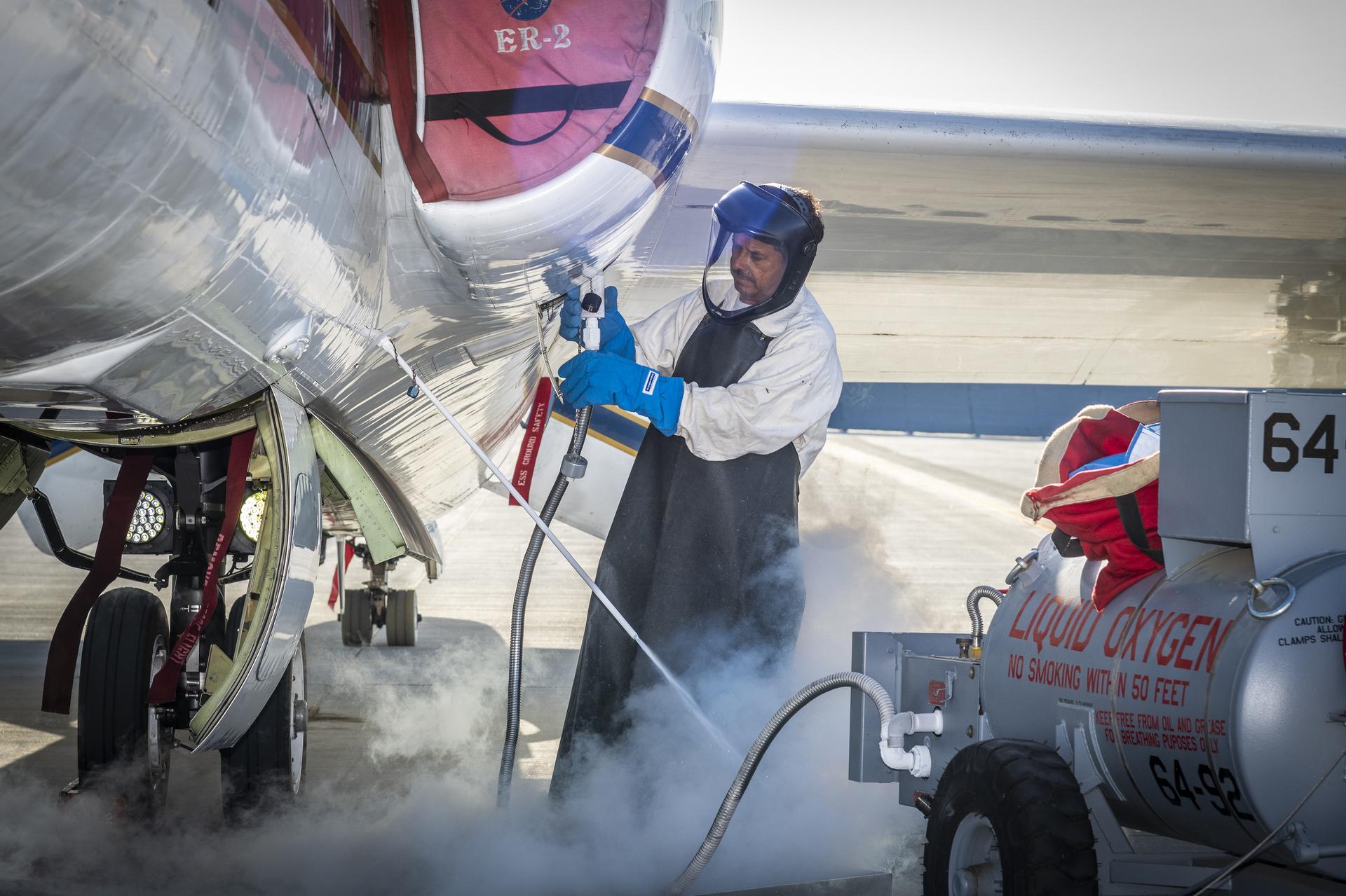 afrc2024 0064 065large.jpg A man clad in a white protective suit, black apron, and blue gloves administers liquid oxygen from a tank to a parked aircraft via hose. Gas clouds hover near his feet and the man is wearing a helmet with a clear visor over his face.