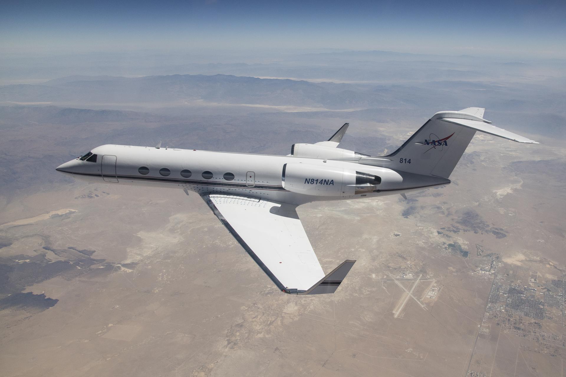 afrc2024 0101 12large.jpg A white Gulfstream IV airplane flies to the left of the frame over a tan desert landscape below and blue mountain ranges in the back of the image. The plane’s tail features the NASA logo, and its wings have winglets. Visible in the lower right third of the image, directly behind the airplane’s wingtip is the Mojave Air and Space Port in Mojave, California.