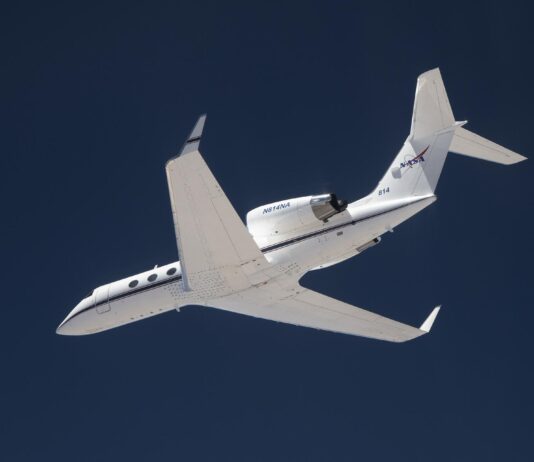 NASA Investigates Earth Science via Innovative Navigation System A white plane flies overhead against a dark blue sky. The plane is heading toward the left of the frame, and the NASA insignia is visible on its tail.